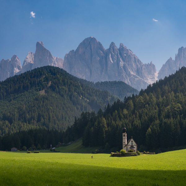 Kleine Kirche auf grüner Wiese vor Wald, dahinter die Dolomiten